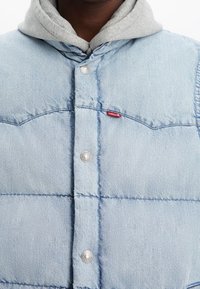 Giacca trapuntata in denim azzurro chiaro con cappuccio grigio, dotata di bottoni a pressione, texture trapuntata e una piccola etichetta rossa del marchio.