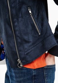 Giacca blu navy con texture in suede, zip argentate, tasche laterali e polsini a coste, indossata sopra una camicia arancione.