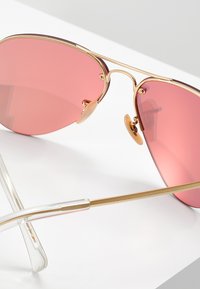 Óculos de sol estilo aviador com armação em metal dourado, lentes com tonalidade rosa e hastes transparentes. As almofadas nasais ajustáveis garantem um ajuste seguro.