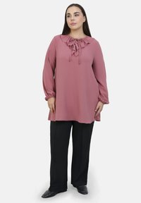 Blusa rosa a maniche lunghe con scollatura arricciata e dettaglio con fiocco, abbinata a pantaloni neri a gamba larga. Tessuto morbido, vestibilità comoda.