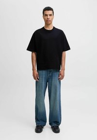 Fekete oversized póló, rövid ujjakkal, széles lábú kék farmernadrággal párosítva. Fekete cipők sima textúrával. Semleges háttér.