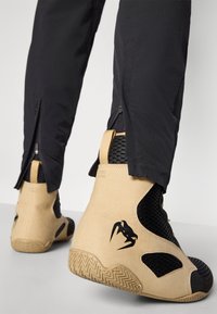 Beige high-top shoes with black mesh accents, textured rubber sole, and a distinctive logo. Zippered pant cuffs reveal shoe details.