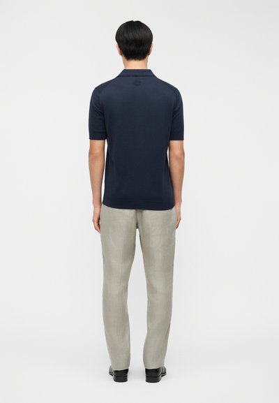 Πόλο μπλουζάκι σε σκούρο μπλε με κοντά μανίκια και κλασικό γιακά, συνδυασμένο με ανοιχτό γκρι παντελόνι από λινό και μαύρα παπούτσια. Εμφανίζεται η πίσω όψη.