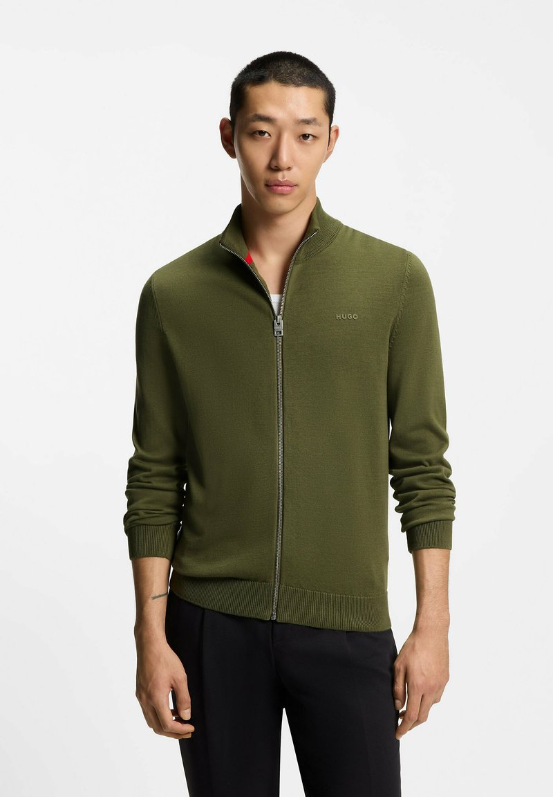 Maglione verde in maglia con zip, maniche lunghe, collo alto e polsini a coste. Logo sul petto e dettaglio contrastante all'interno del collo.