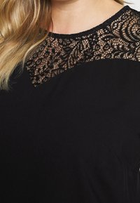 Sort top med en gennemsigtig blonde-udskæring med indviklede blomster mønstre. Glat, blødt stof med en afslappet pasform og rund krave.