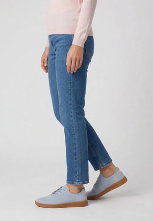 Osoba ubrana w jasnoniebieskie dżinsy, bladoróżowy sweter i jasnoszare trampki z brązowymi podeszwami stoi na neutralnym tle.