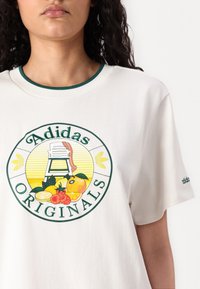 Balta medvilninė marškinėlių su apvaliu grafiku, kuriame pavaizduota kėdė ir įvairūs vaisiai, žali akcentai ant apykaklės ir prekės ženklo logotipas ant rankovės.