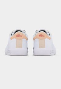 Ζεύγος λευκών αθλητικών παπουτσιών με ταμπάλες τακουνιού σε ανοιχτό μπεζ και ροζ λογότυπα κροκοδείλου, φωτογραφημένα από πίσω σε απλό ανοιχτό φόντο.