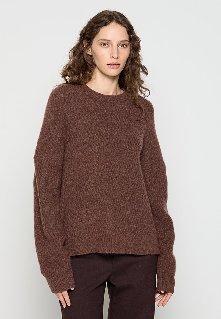 Brauner strukturierter Pullover mit Zickzackmuster, rundem Halsausschnitt und langen Ärmeln, aus einem weichen, kuscheligen Material gefertigt. Kombiniert mit dunklen Hosen.