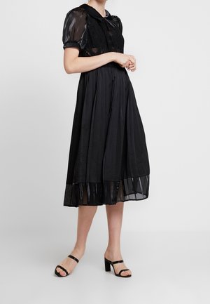 Femme portant une robe midi noire avec des manches bouffantes transparentes, un corsage en dentelle, une jupe plissée, un ourlet transparent et des sandales à talons noires.