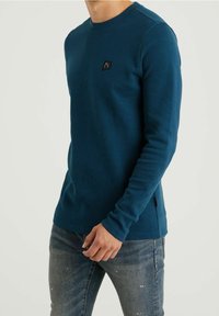 Blå långärmad skjorta med struktur i våffelstickning, med en liten svart logotyp på bröstet och åtsittande ärmslut.