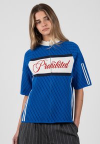 Mujer joven con cabello largo que lleva una camiseta azul de manga corta con cremallera con la palabra "Prohibido" en letras rojas sobre el pecho y pantalones grises a rayas.