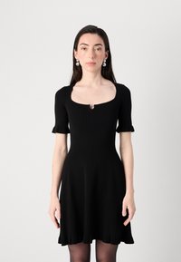 A model stands facing the camera, wearing a fitted black ribbed dress with short sleeves and a keyhole detail, accessorized with silver earrings.