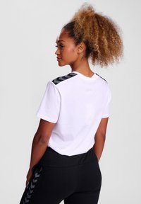 Hvid t-shirt med korte ærmer, prydet med sorte chevron-mønstrede detaljer på skuldrene. Bæres med sorte bukser, hvor den teksturerede stof er synlig.