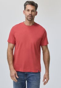 Homme portant un t-shirt rouge uni à manches courtes et un jean bleu, debout devant un fond gris clair.
