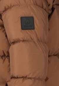 Braune Pufferjacke mit auffälliger steppierter Textur am Ärmel, versehen mit einem schwarzen Logo-Patch mit geometrischem Design an der Armseite.
