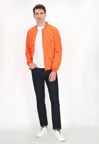 Giacca bomber arancione con motivo floreale, abbinata a una t-shirt bianca, jeans scuri e sneakers bianche. Design semplice e casual.