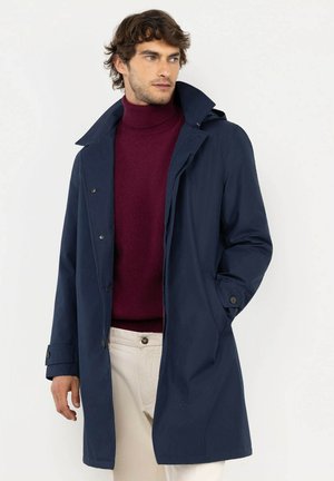 Hombre con abrigo azul marino sobre un suéter de cuello alto burdeos y pantalones beige, mirando hacia un lado con una mano en el bolsillo del abrigo.
