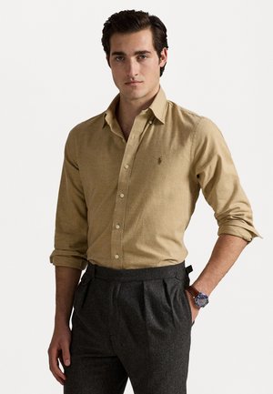 Cămașă bej cu nasturi și mâneci lungi, din țesătură texturată, având un mic logo. Purtați cu pantaloni gri pliați și un ceas albastru la încheietura mâinii.