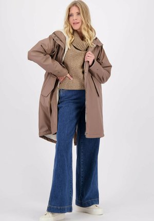 Blonde vrouw in een tan gewatteerde jas over een bruine coltrui, met een brede blauwe spijkerbroek en witte platform schoenen, staand tegen een witte achtergrond.