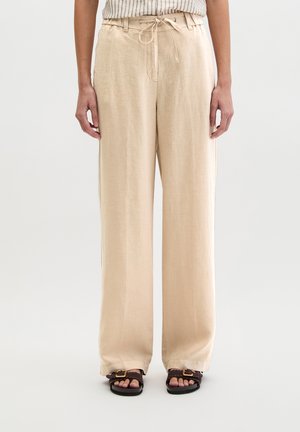 Frau trägt beigefarbene Leinenhose mit weitem Bein und Kordelzugbund sowie dunkelbraune Sandalen mit Schnalle, steht vor weißem Hintergrund.