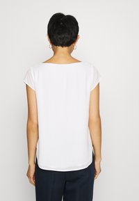 Blouse blanche à manches courtes avec un large col, texture lisse et ourlet légèrement incurvé, vue de dos. Design simple et classique.