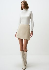 Cremiger Rollkragenpullover kombiniert mit einem beigefarbenen Miniröckchen. Das Model trägt knielange, glänzende weiße Stiefel. Glattes Material und schlichtes Design.