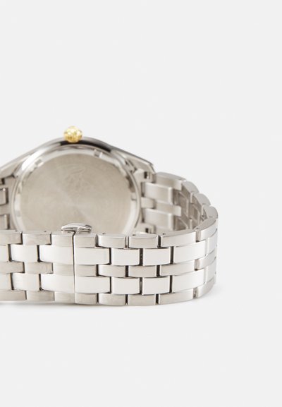 Montre-bracelet en métal argenté avec un boîtier rond lisse, un dos texturé et une couronne dorée. Le bracelet est composé de maillons rectangulaires reliés.