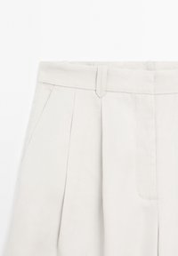 Pantalon beige taille haute avec plis à l'avant, passants de ceinture et poches latérales, visible de la taille jusqu'à mi-cuisse.