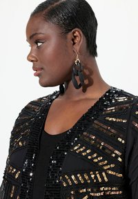 Cárdigan negro adornado con lentejuelas negras y doradas en patrones geométricos. Presenta un escote en V profundo y se lleva sobre una blusa negra. Pendientes negros.