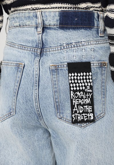 Jeans in denim blu sbiadito con tasche posteriori, cuciture decorative e un grande etichetta nera con testo bianco: "EROISMO E ROYALTY DELLE STRADE."