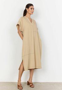 Beige jurk tot de knie met een V-hals, korte mouwen, zijsplitten en zichtbare stiksels. Draagt met bruine blokhak sandalen.