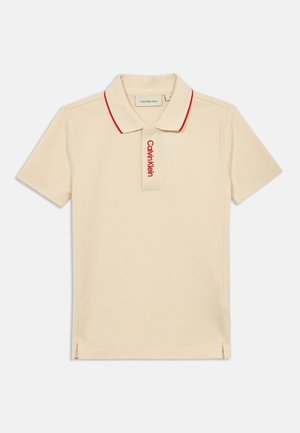 Polo bege com colarinho vermelho e logótipo bordado "Calvin Klein" no lado esquerdo do peito. Mangas curtas, aberturas laterais na bainha.