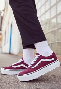 Sneakers in suede bordeaux con strisce e lacci bianchi, suola spessa bianca, indossate con calzini bianchi alti e pantaloni neri.