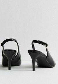 Escarpins noirs brillants à bride arrière avec un bout pointu, un talon aiguille fin et une sangle de cheville réglable. Texture lisse sans motifs visibles.
