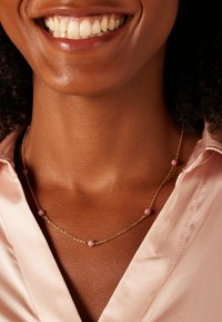 Collier en chaîne dorée avec des perles roses, design délicat, texture lisse, porté contre une chemise en satin rose, mettant en valeur le décolleté et les bijoux.