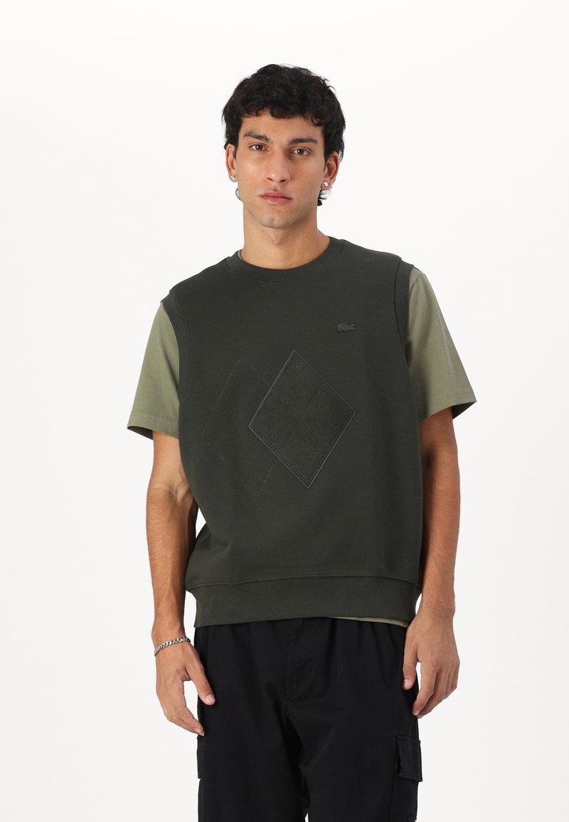 Felpa senza maniche verde scuro con design a diamante testurizzato e logo, abbinata a una camicia oliva a maniche corte sotto. Materiale in misto cotone.