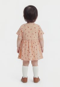 Copil mic văzut din spate, purtând o rochie cu imprimeu floral piersică, șosete albe până la genunchi și pantofi maro, pe un fundal alb simplu.