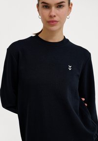 Jonge vrouw draagt een zwart shirt met lange mouwen en ronde hals met klein wit dubbel-chevron logo op de linkerborst, staand tegen een effen witte achtergrond.