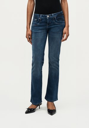 Moteris dėvi tamsiai mėlynas išsiplėtusias džinsus ir juodus batelius su smailiais pirštais ir žemu kulniuku, stovinti prie paprasto šviesaus fono.