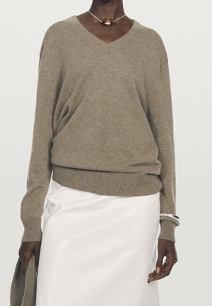 Personne portant un pull taupe à col en V et une jupe blanche, tenant un tissu taupe assorti et portant un collier pendentif rond ainsi qu'un bracelet en argent.
