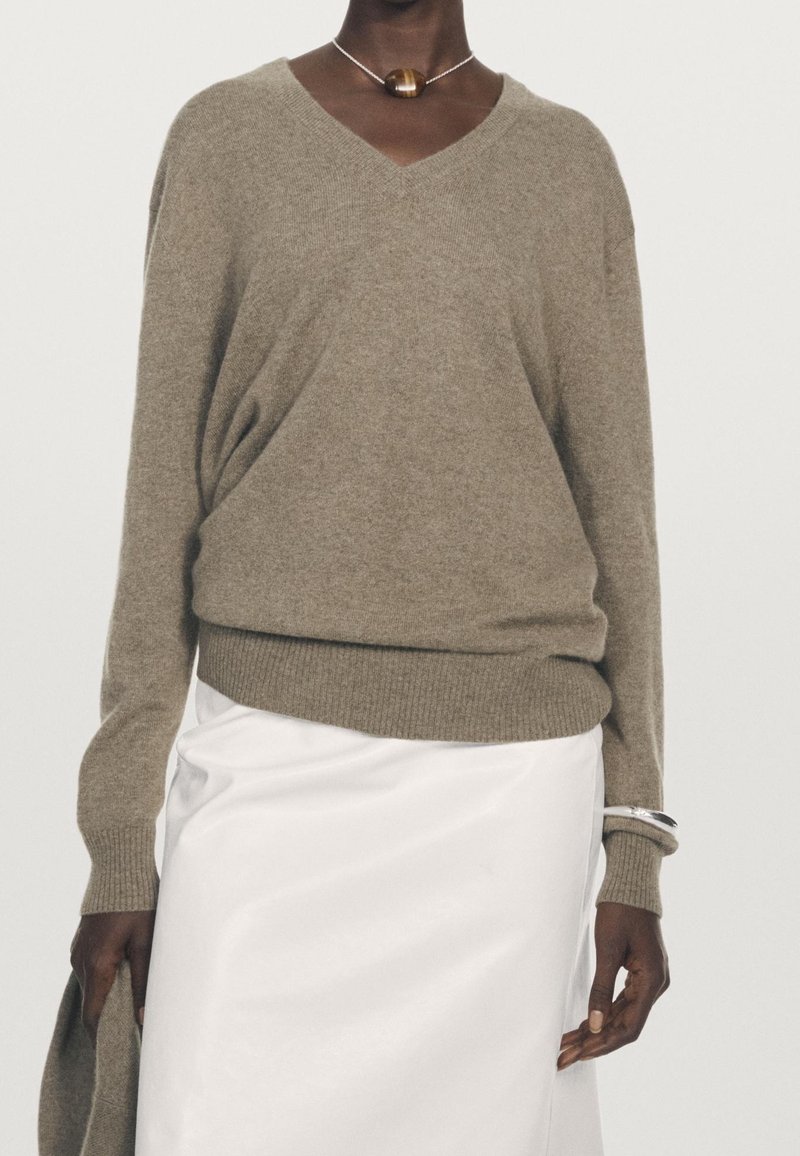 Personne portant un pull taupe à col en V et une jupe blanche, tenant un tissu taupe assorti et portant un collier pendentif rond ainsi qu'un bracelet en argent.