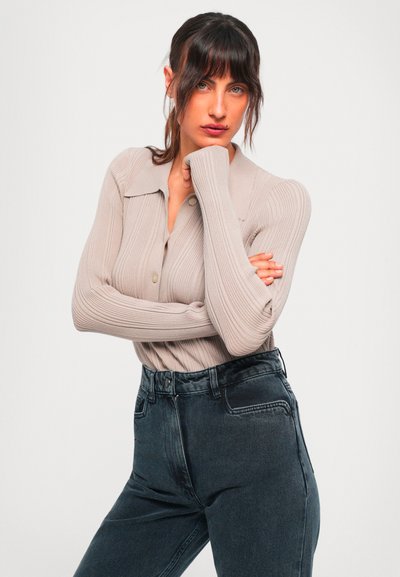 Femeie cu părul închis la culoare, purtând un top lung cu mâneci raglan de un bej deschis cu dungi și blugi închiși la culoare cu talie înaltă, pozând cu o mână lângă bărbie.