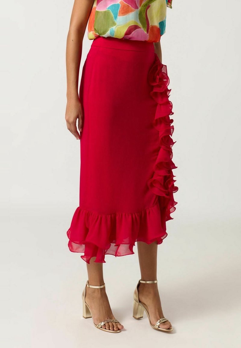 Femme portant une jupe mi-longue rouge vif avec des détails volantés sur un côté et à l'ourlet, associée à des sandales à talons hauts dorées.