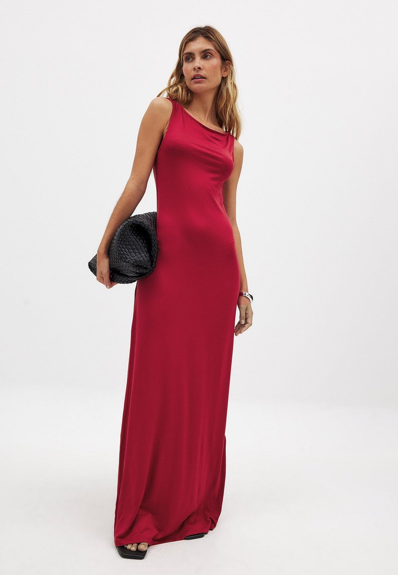 Robe longue sans manches de couleur rouge avec un décolleté drapé, texture lisse et silhouette ajustée, accompagnée d'une pochette tissée noire.