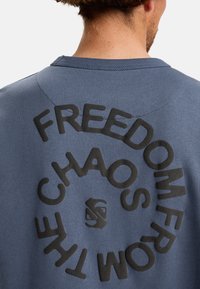 Felpa blu con colletto a girocollo, caratterizzata da un audace design di testo nero sul retro: "LIBERTÀ DAL CAOS." Tessuto morbido.