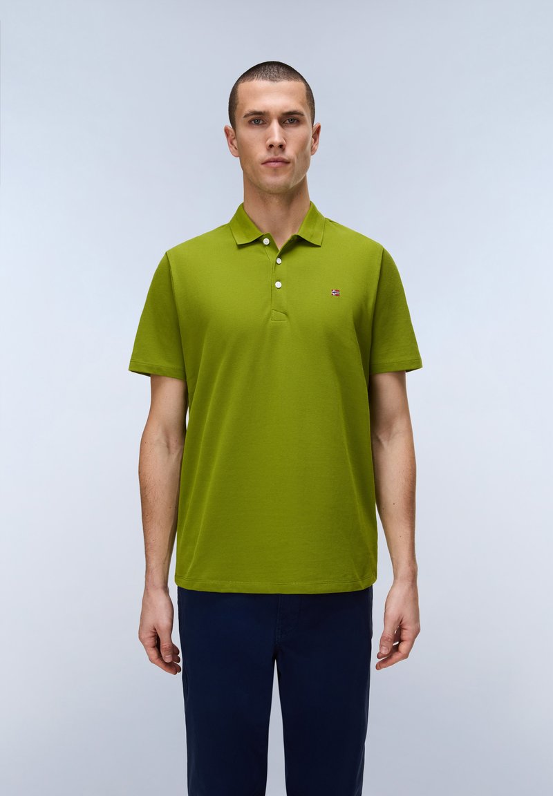 Männliches Model mit kurzen Haaren, das ein grünes Poloshirt mit kurzen Ärmeln und dunkelblaue Hosen trägt und vor einem schlichten hellen Hintergrund steht.