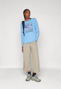 Light blue long-sleeve shirt with graphic design featuring mountains, paired with beige wide-leg pants and gray hiking shoes. Backpack on shoulder.