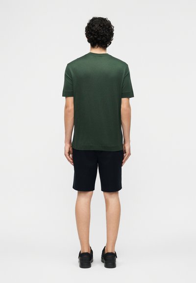 Άτομο με σγουρά μαλλιά που φοράει σκούρο πράσινο κοντομάνικο μπλουζάκι, μαύρο σορτς και μαύρα παπούτσια, στέκεται με την πλάτη προς τα εμπρός σε λευκό φόντο.