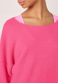 Pull rose avec une texture douce, un large décolleté et des épaules tombantes, superposé à un débardeur rose.